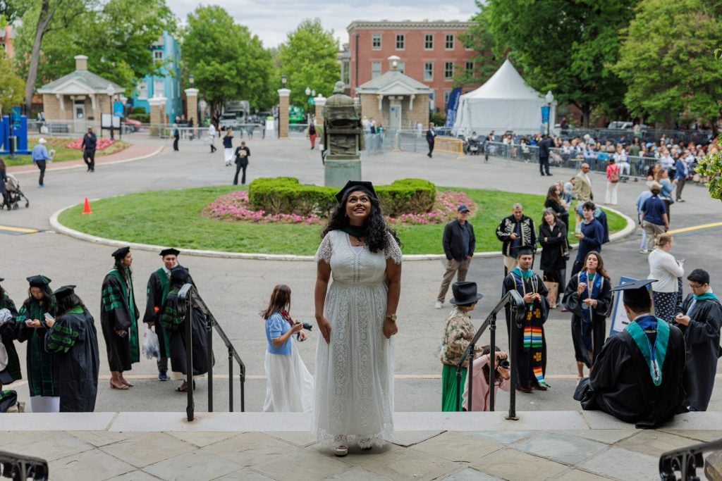 A female student in a graduation cap and white dress looks up at a building and smiles in wonder