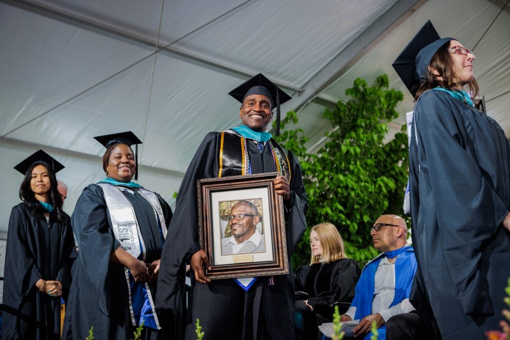 A male student in a cap and gown smiles while carrying a framed photo of a male relative