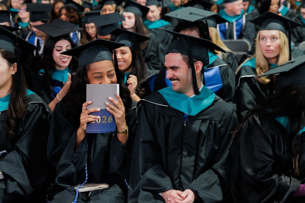 A female student holds up a program  book to her face and smiles while a male student sitting next to her laughs. 