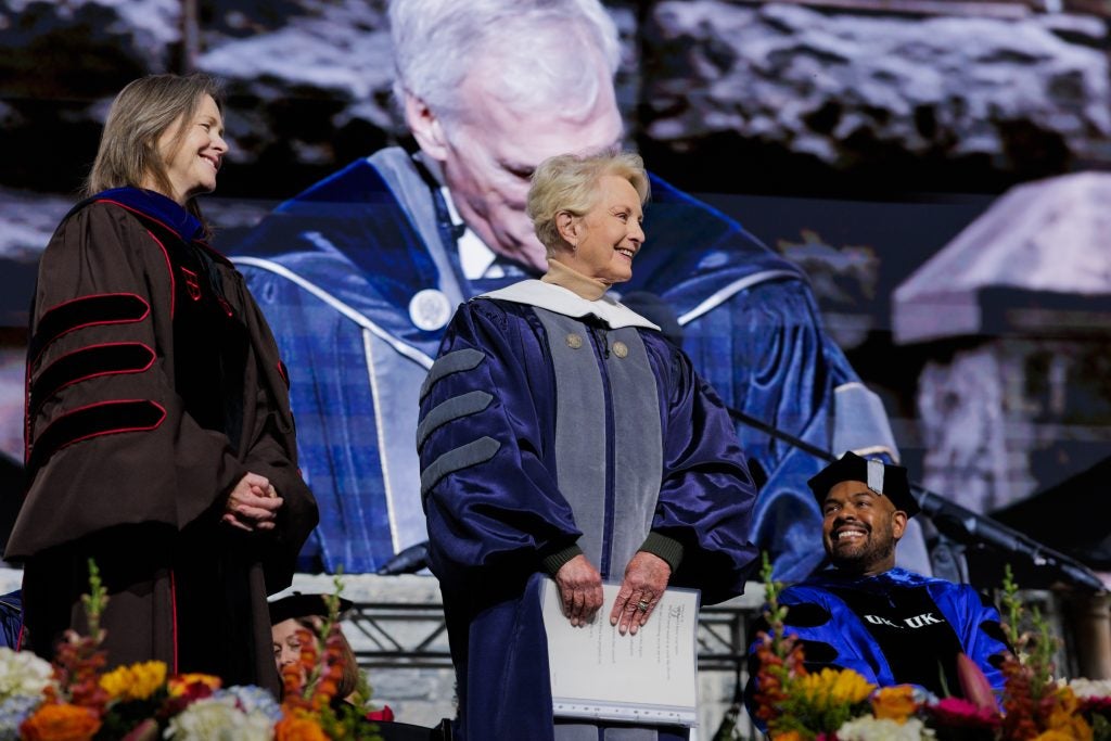 Cindy McCain smiles onstage in a navy graduation gown.