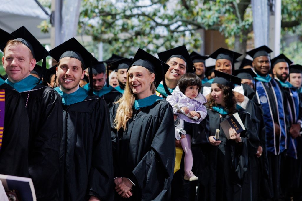 A group of students in graduation cap and gowns smile in a line. One student holds a baby