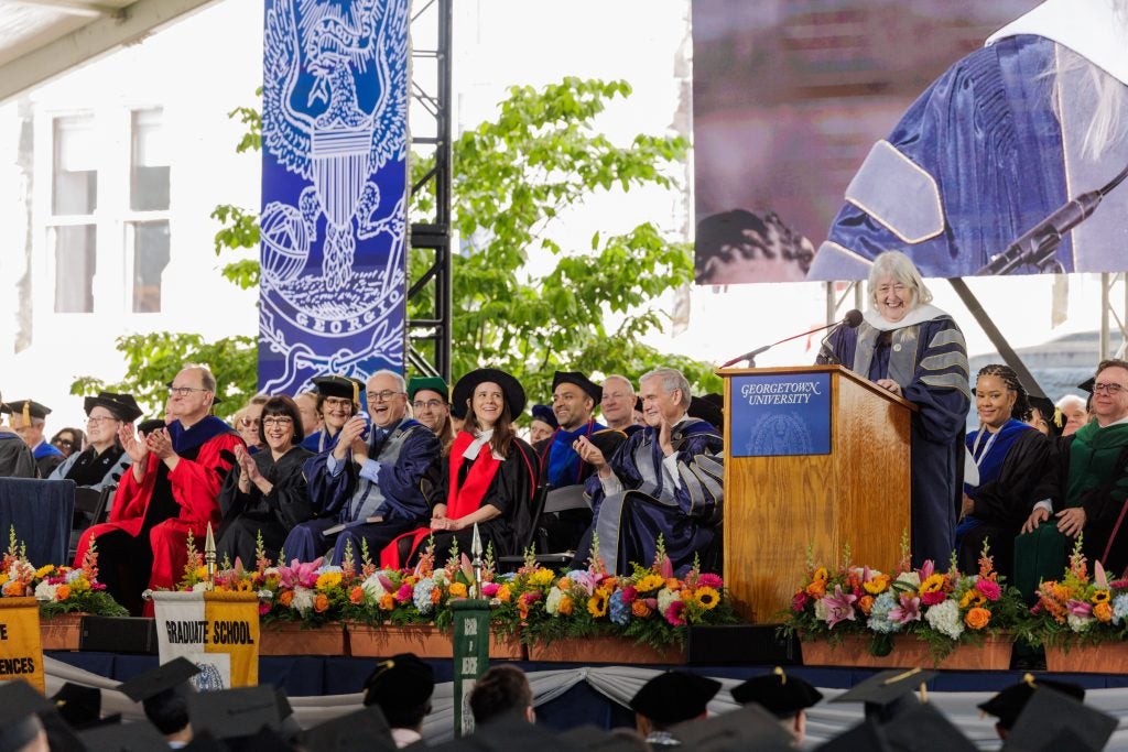 Older Caucasian woman in commencement regalia speaks to a crowd