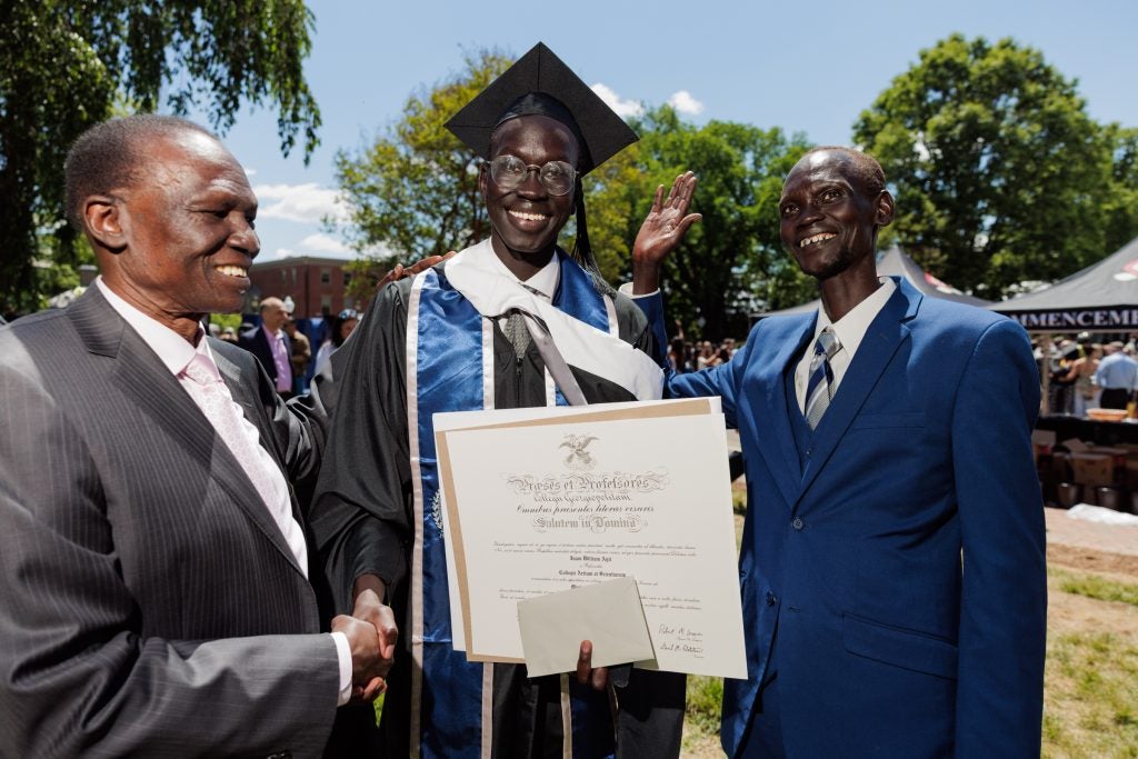 A male student in a graduation cap and gown smiles while standing alongside his relatives