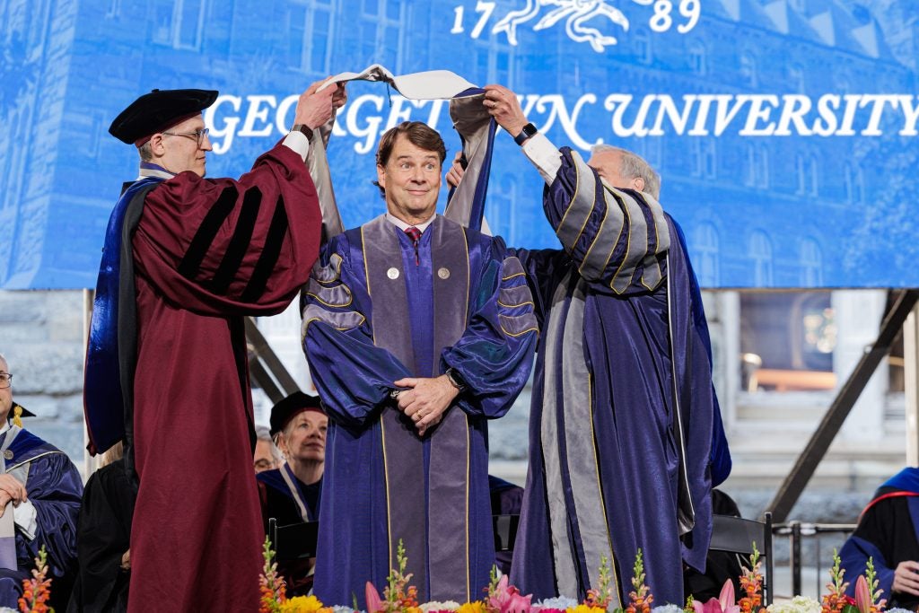 Two men place a sash over another man in a purple graduation gown