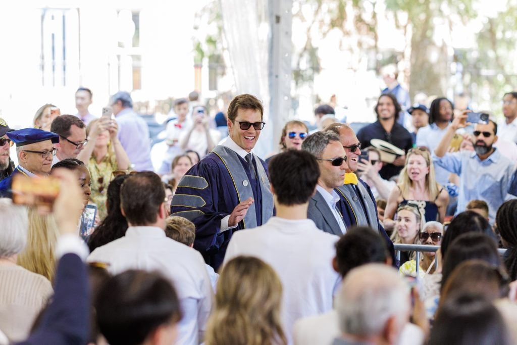 Tom Brady wears a purple graduation robe and sunglasses and waves as he walks through a crowd
