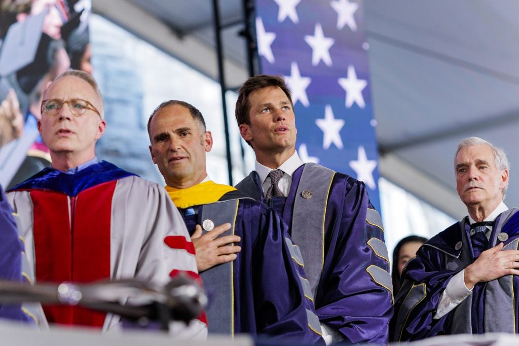 Tom Brady sings the national anthem  in front of an American flag. He wears a purple graduation robe