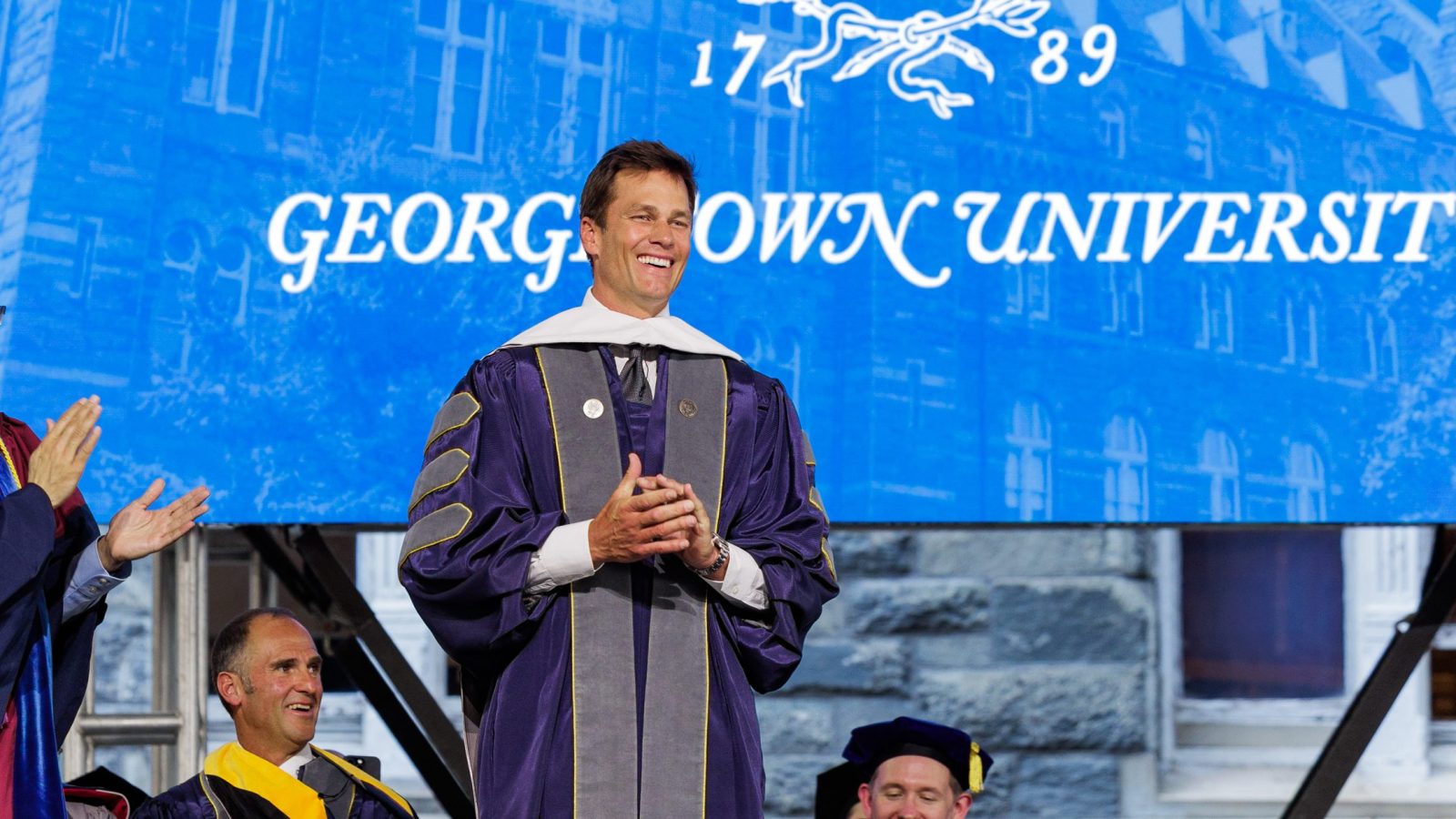 Tom Brady smiles in a purple graduation robe in front of a blue screen that says "Georgetown University"