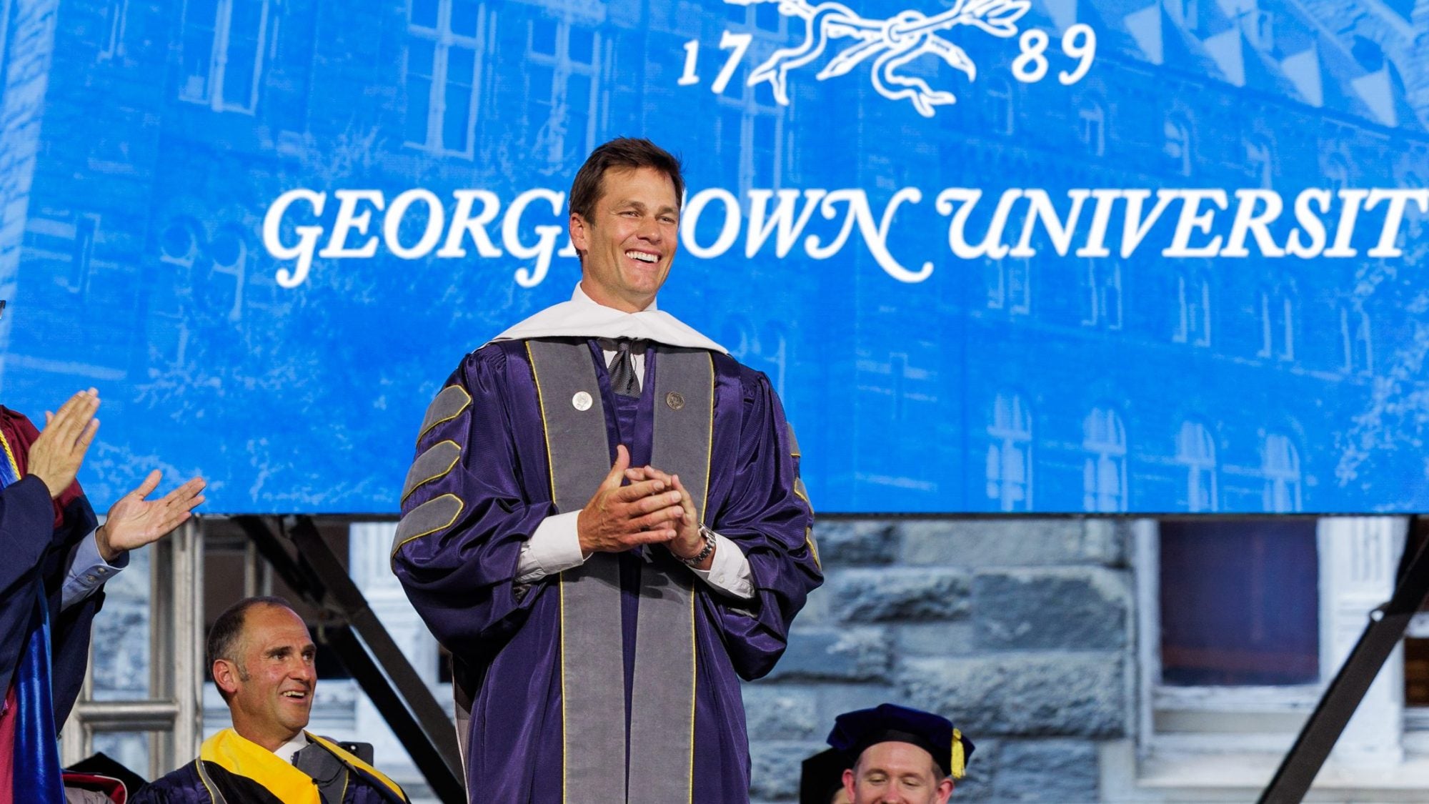 Tom Brady smiles in a purple graduation robe in front of a blue screen that says "Georgetown University"