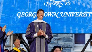 Tom Brady smiles in a purple graduation robe in front of a blue screen that says "Georgetown University"