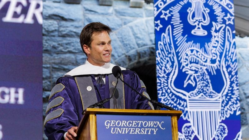 Tom Brady stands behind a podium in a purple graduation gown