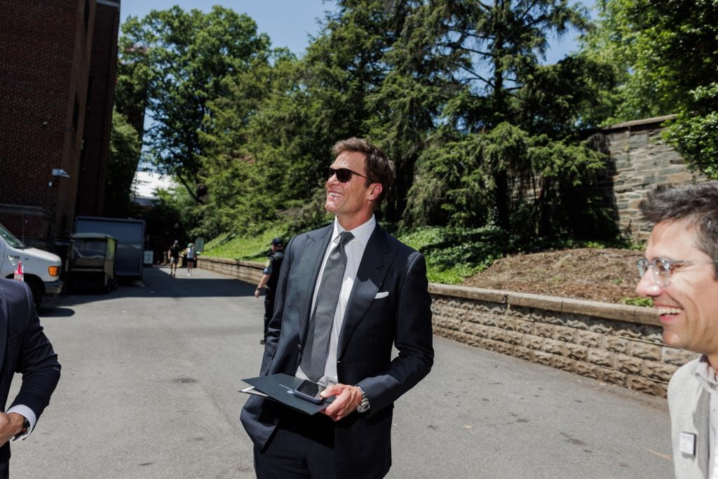 Tom Brady smiles on a sunny day. He wears sunglasses and a suit and tie