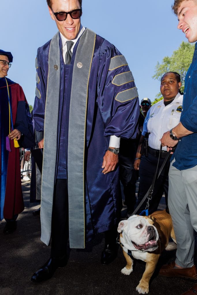 Tom Brady pets a dog while wearing a purple graduation gown
