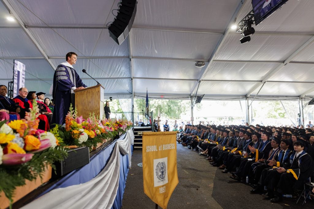 Tom Brady delivers a speech onstage from behind a podium in front of graduating seniors