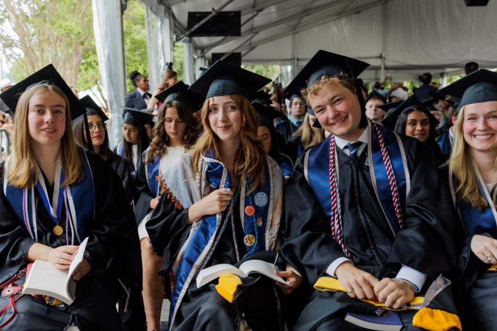 Three students smile in graduation gowns while sitting in an audience