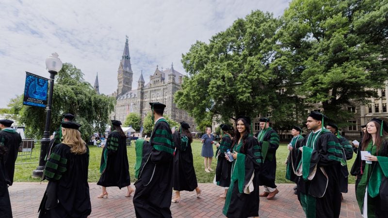 A group of students in green and black graduation gowns walk on Georgetown's campus