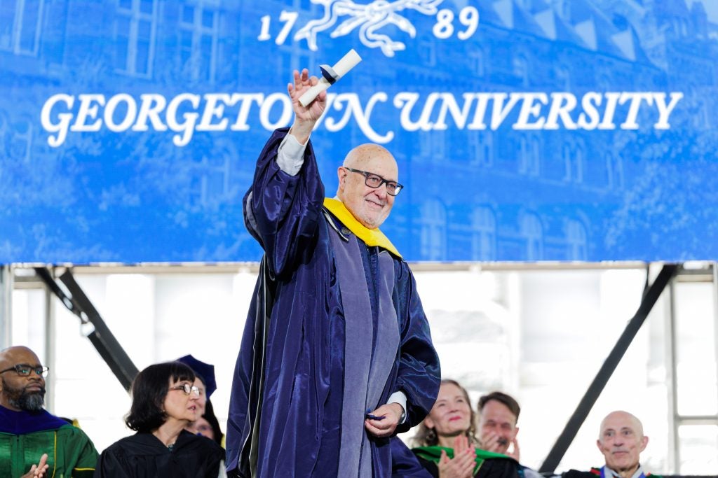 Older Caucasian man holds up a wrapped diploma on stage while in grad gown