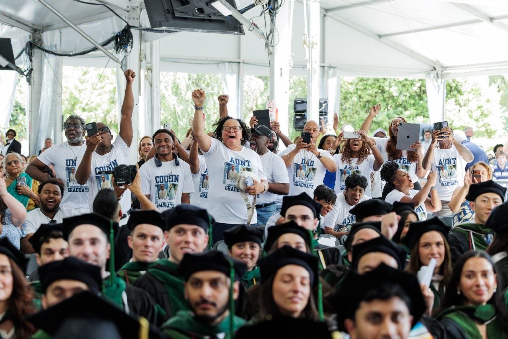 A family jumps and cheers during a graduation ceremony. They each wear T-shirts designating their relationship to the graduate