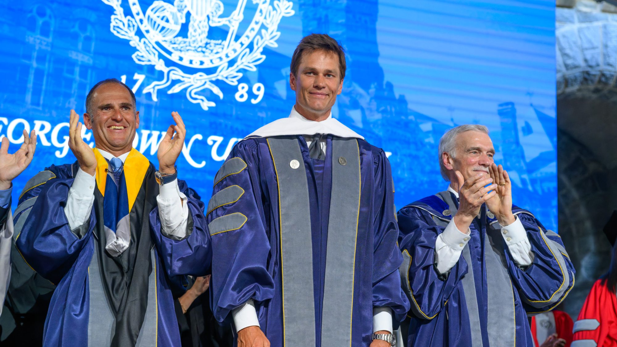 Tom Brady smiling flanked by two other men clapping on a graduation stage