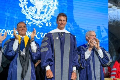 Tom Brady smiling flanked by two other men clapping on a graduation stage