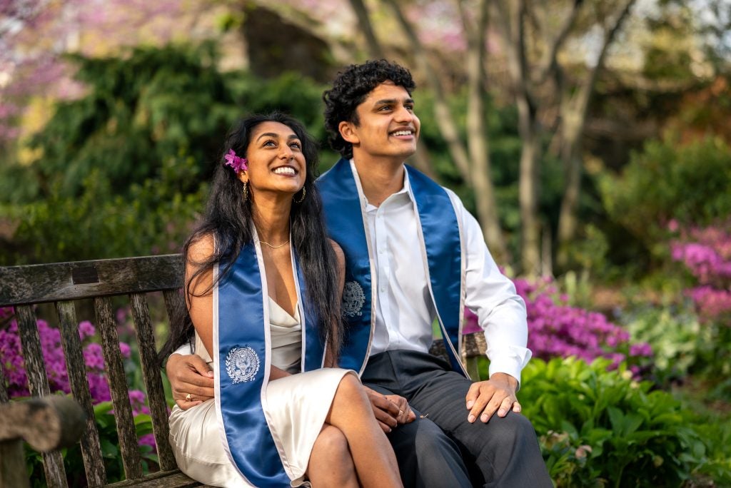 Two students with hands around each other wearing white attire and graduation stoles in a garden