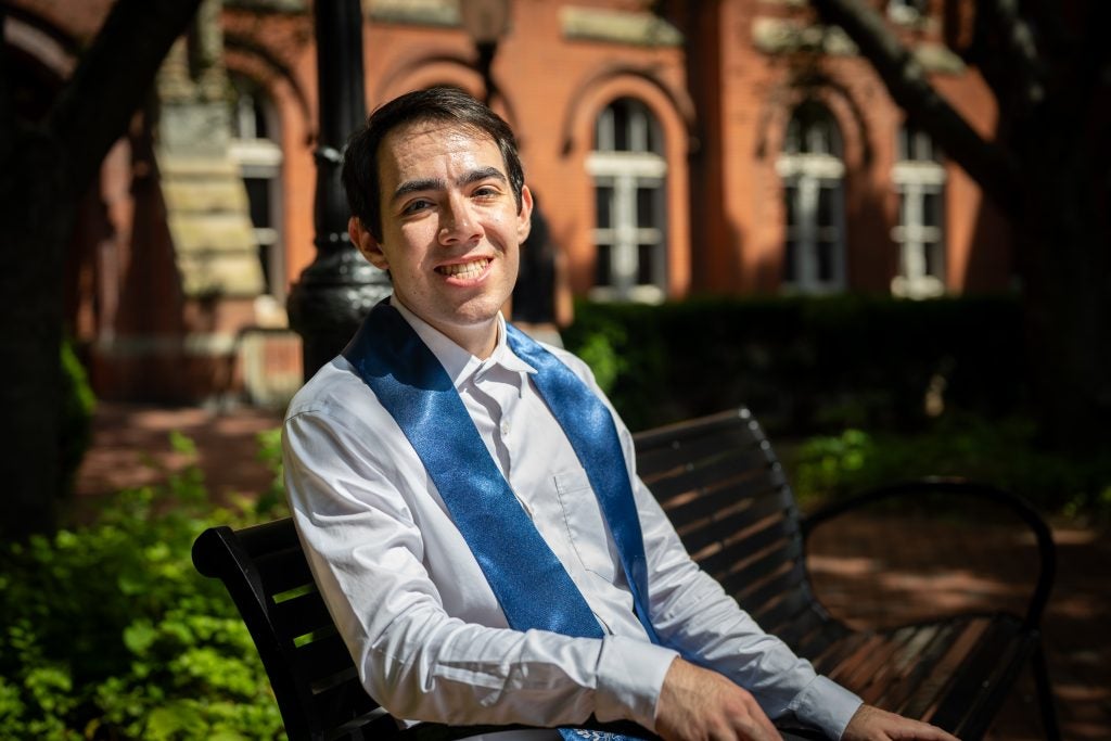 Young man in white shirt with a graduation stole sitting on a bench and smiling