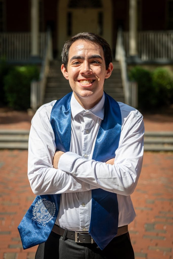 Young man in white shirt with a graduation stole with arms crossed and smiling on a sunny day