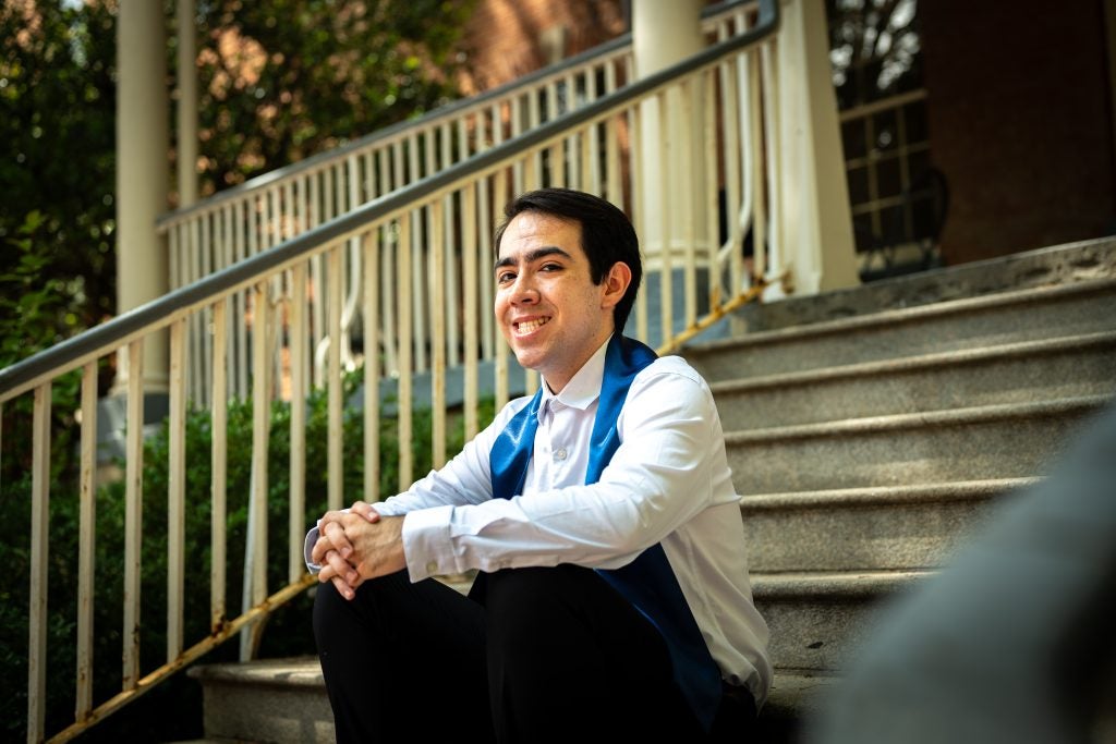 Young man in white shirt with a graduation stole sitting on steps and smiling