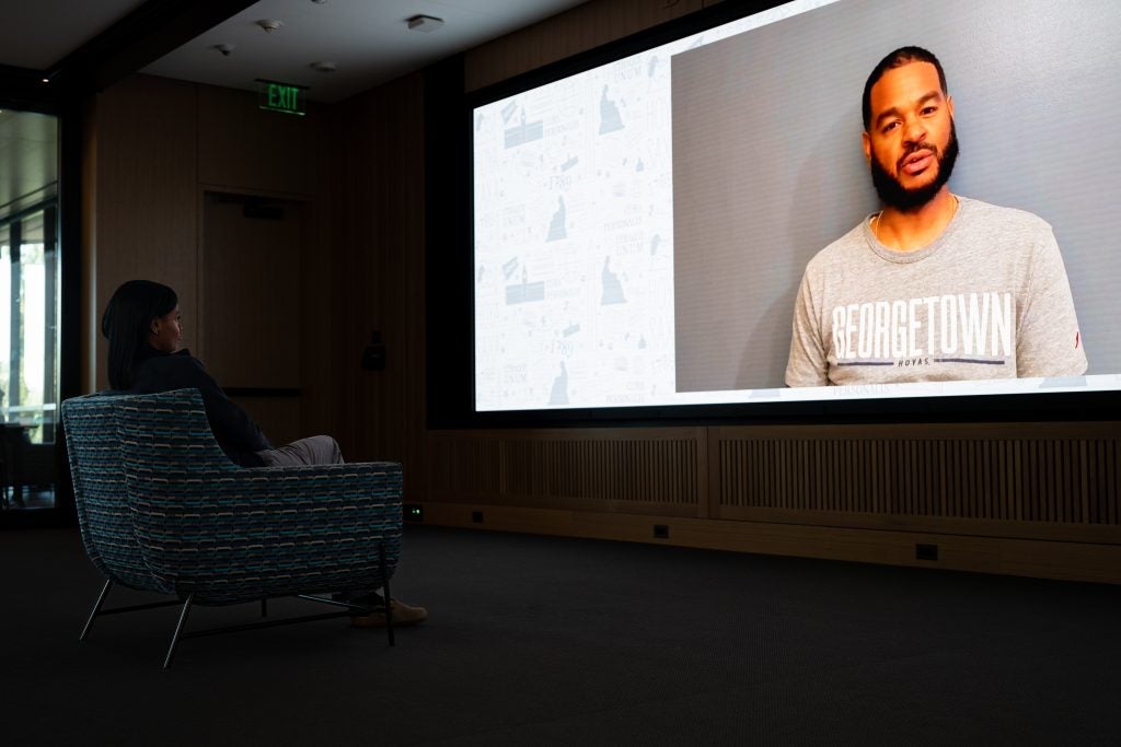 Young Black woman in a dark room watching her dad speak on a big screen