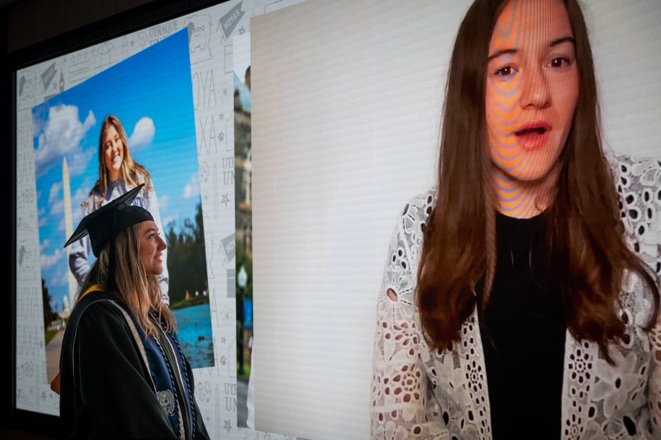 Young woman in graduation regalia looks at an image of her sister on a large screen