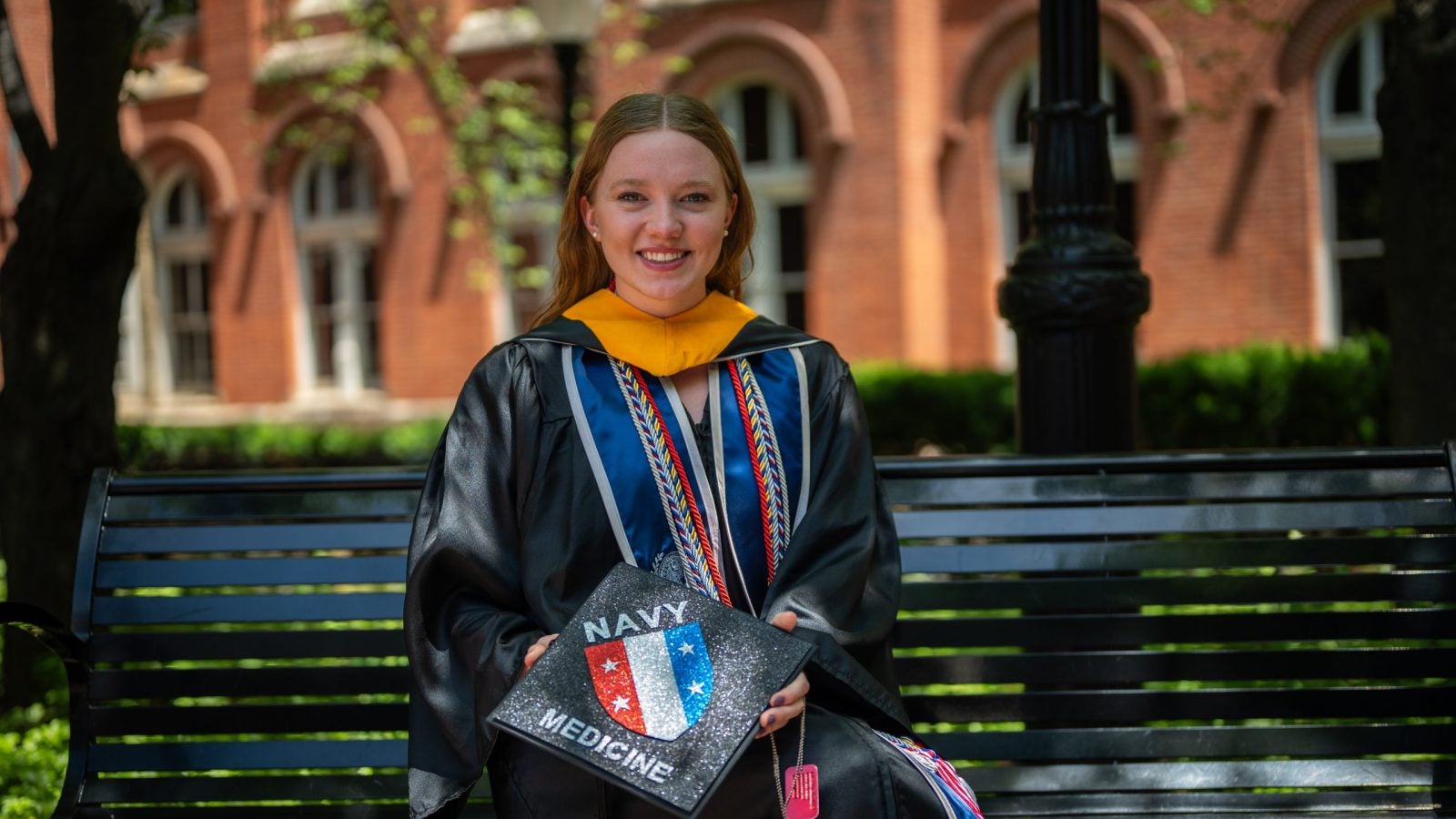 Young Caucasian woman smiling in grad regalia holding a grad cap and sitting on a bench