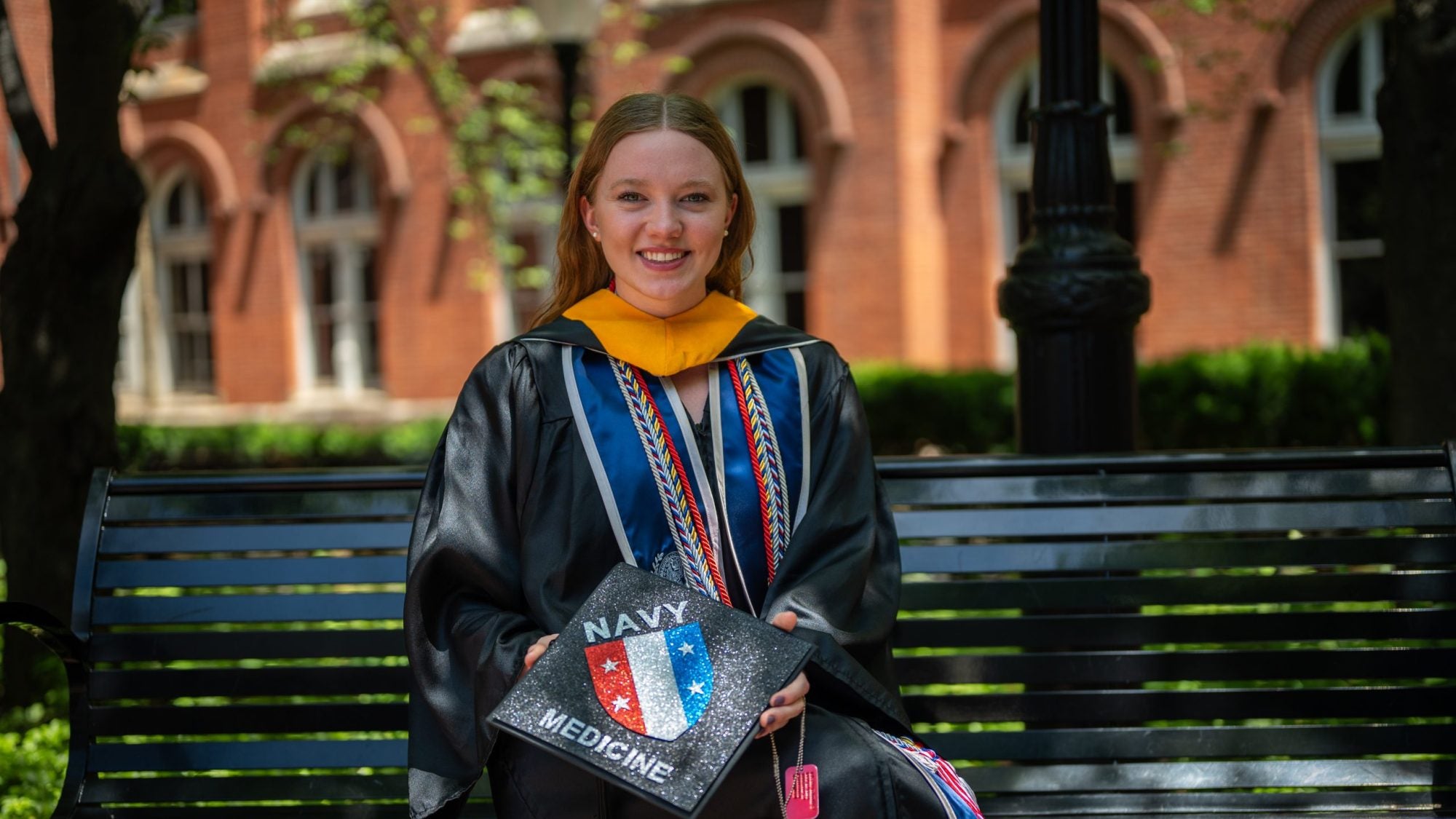 Young Caucasian woman smiling in grad regalia holding a grad cap and sitting on a bench
