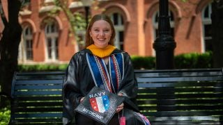 Young Caucasian woman smiling in grad regalia holding a grad cap and sitting on a bench