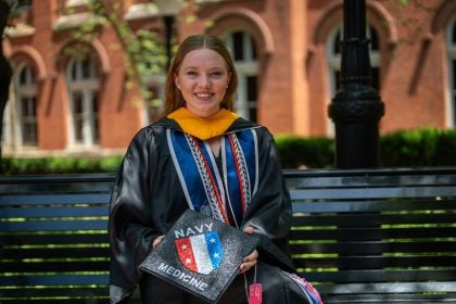 Young Caucasian woman smiling in grad regalia holding a grad cap and sitting on a bench