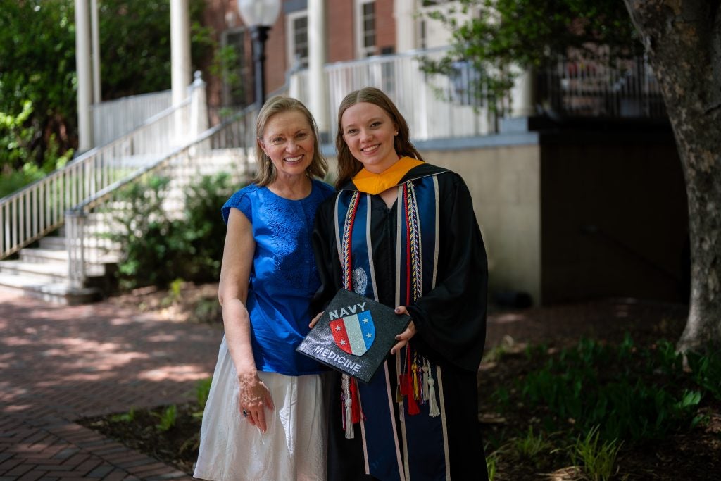 Young Caucasian woman smiling in grad regalia next to her mom while holding a grad cap