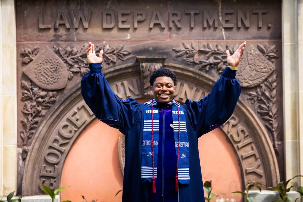 A young holds up both his arms in celebration in front of a plaque that says Georgetown Law. He wears a graduation robe