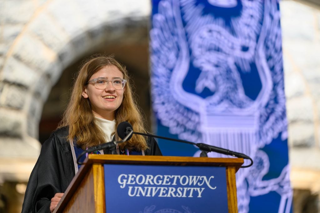 A female student in glasses smiles while speaking behind a podium that says "Georgetown University"