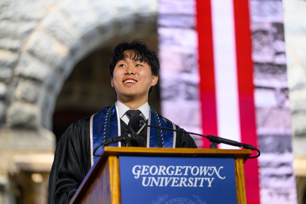 A male student in a graduation gown smiles from behind a podium that says "Georgetown University"