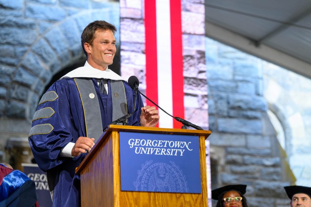 Tom Brady smiling in grad gown while speaking at podium