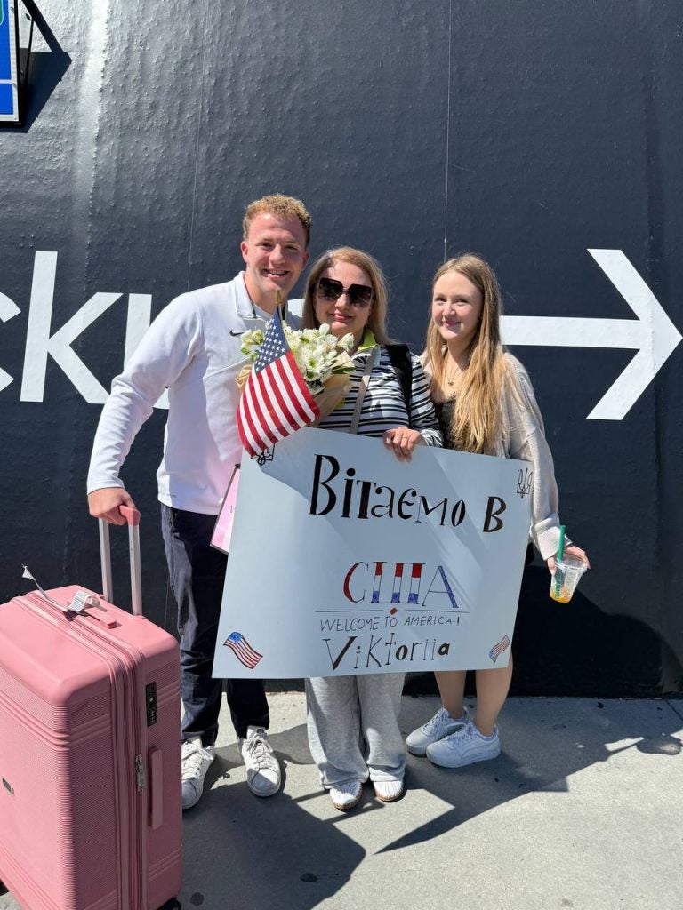 Two young adults with a welcome sign welcoming an older woman
