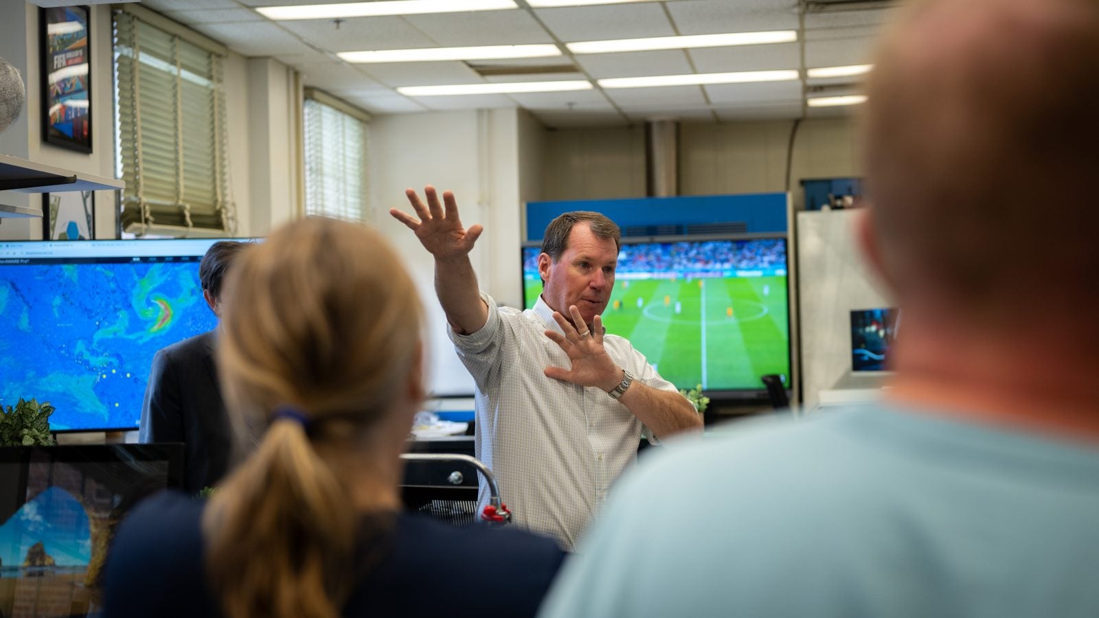 A man makes a gesture with his arms while speaking with a TV behind him playing a soccer match