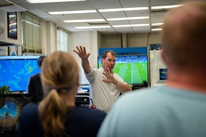 A man makes a gesture with his arms while speaking with a TV behind him playing a soccer match