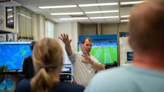 A man makes a gesture with his arms while speaking with a TV behind him playing a soccer match