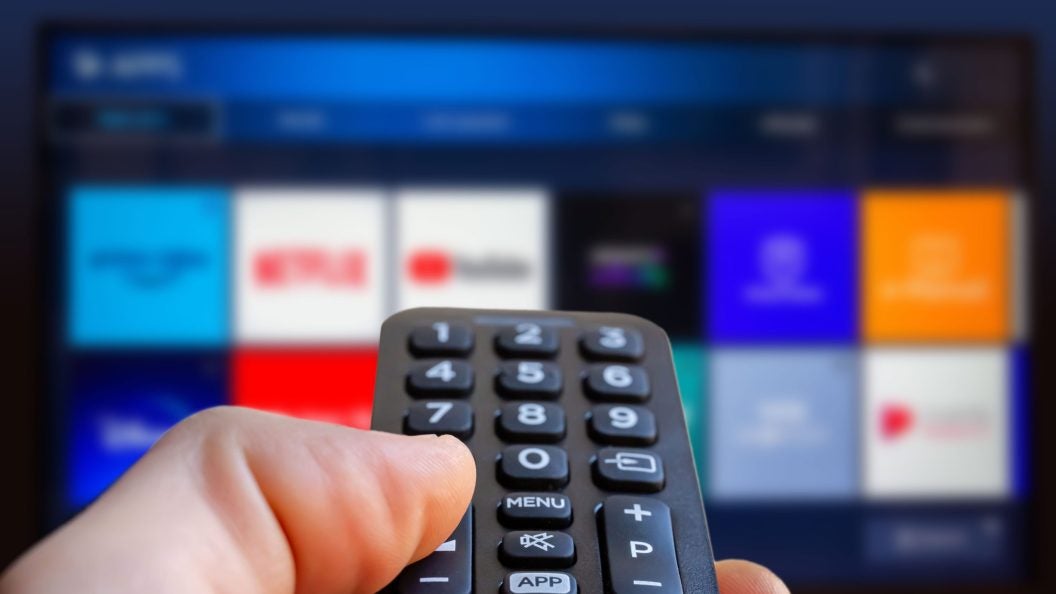 A man is holding a remote control of a smart TV in his hand. In the background you can see the television screen with streaming entertainment apps for video on demand