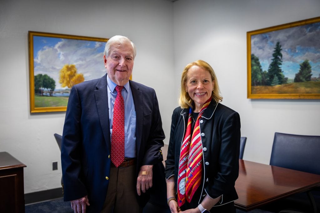 A man with white hair and a suit smiles with a woman in a suit