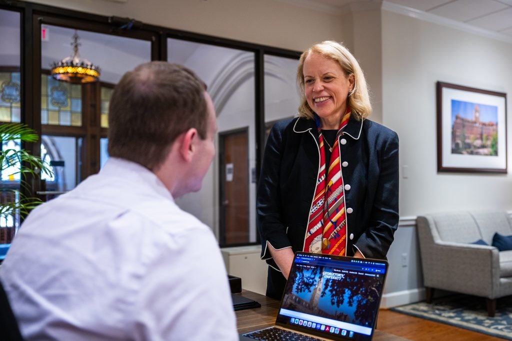 A woman in a navy suit smiles at a colleague sitting behind a desk