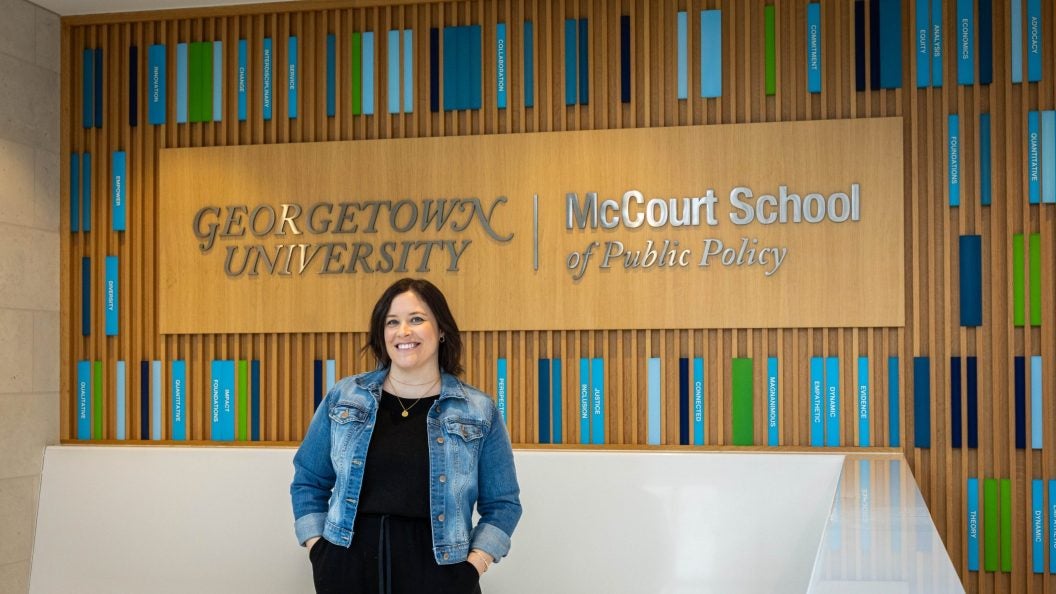 A woman in a jean jacket stands in front of a sign that says "Georgetown University McCourt School of Public Policy"