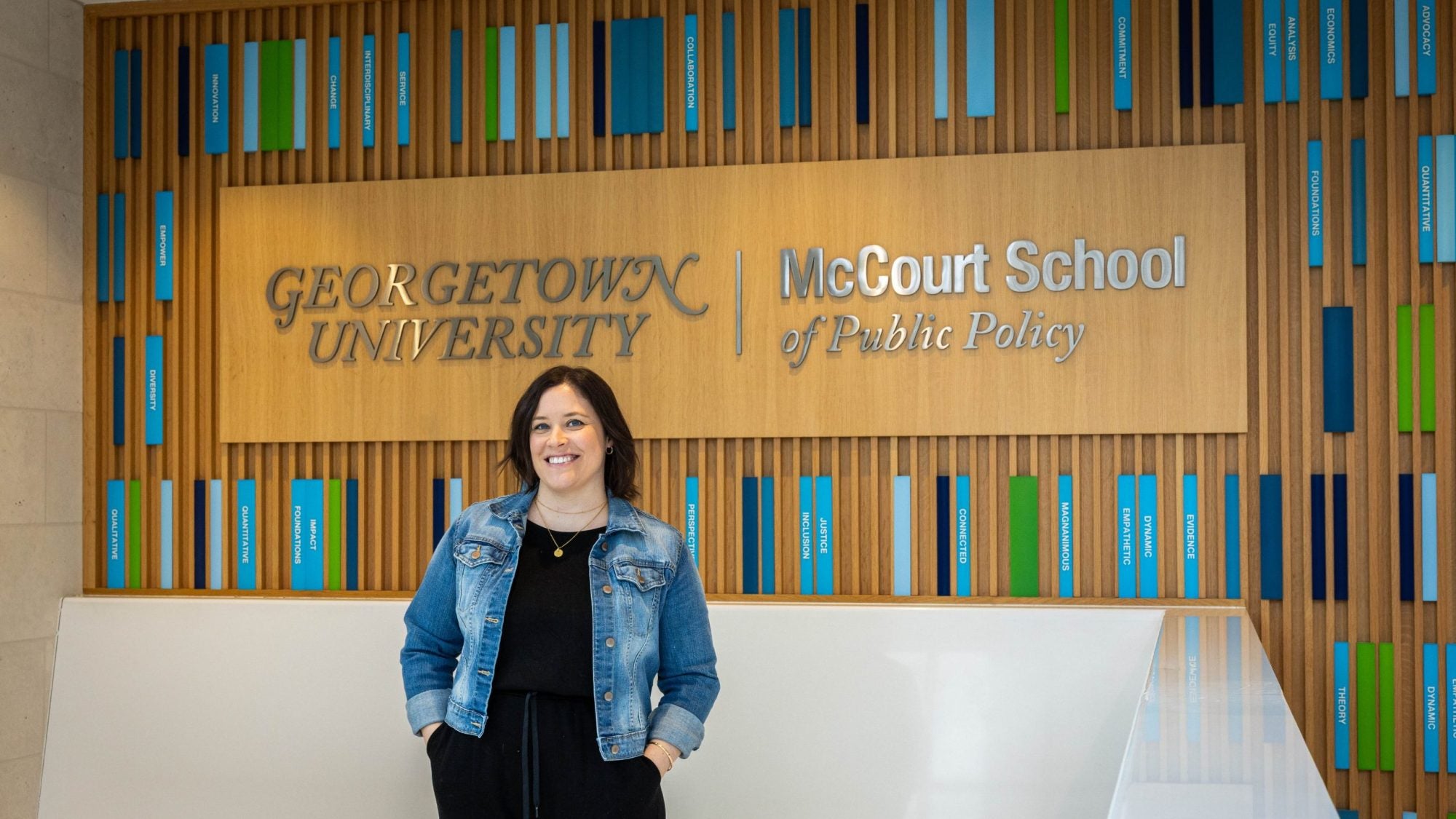 A woman in a jean jacket stands in front of a sign that says "Georgetown University McCourt School of Public Policy"