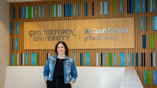 A woman in a jean jacket stands in front of a sign that says "Georgetown University McCourt School of Public Policy"