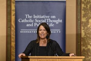A woman with brown hair and a black sweater stands behind a podium speaking