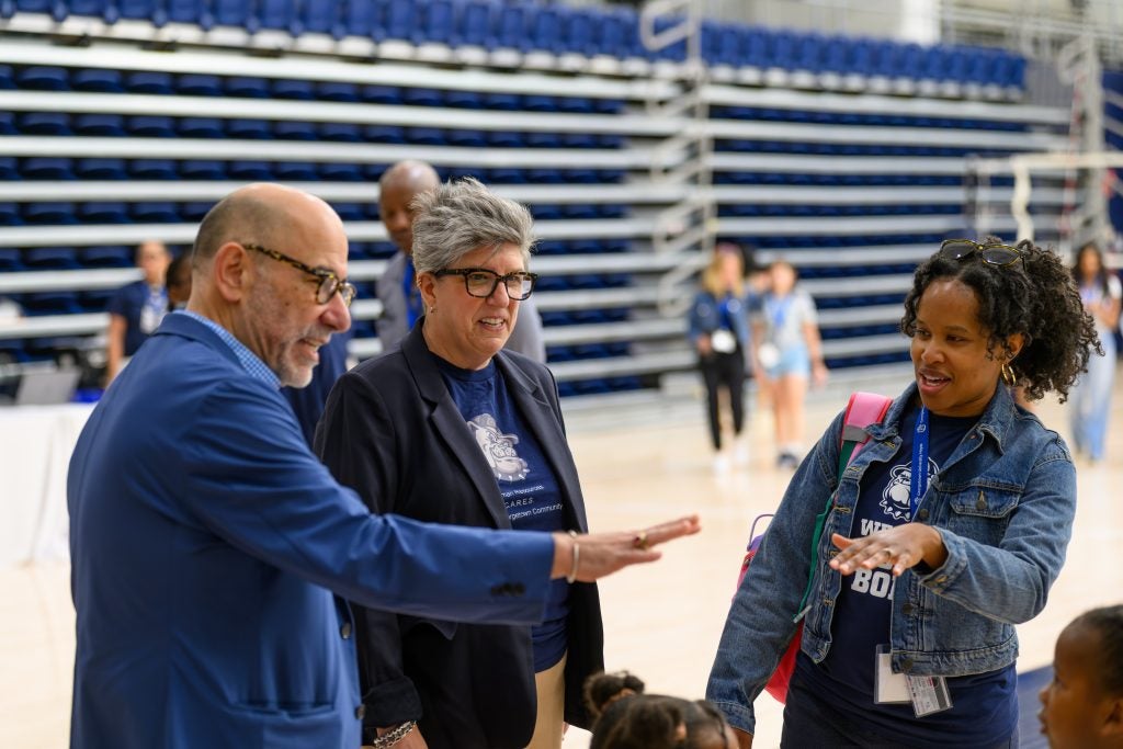 Three people in business attire talk in a gym